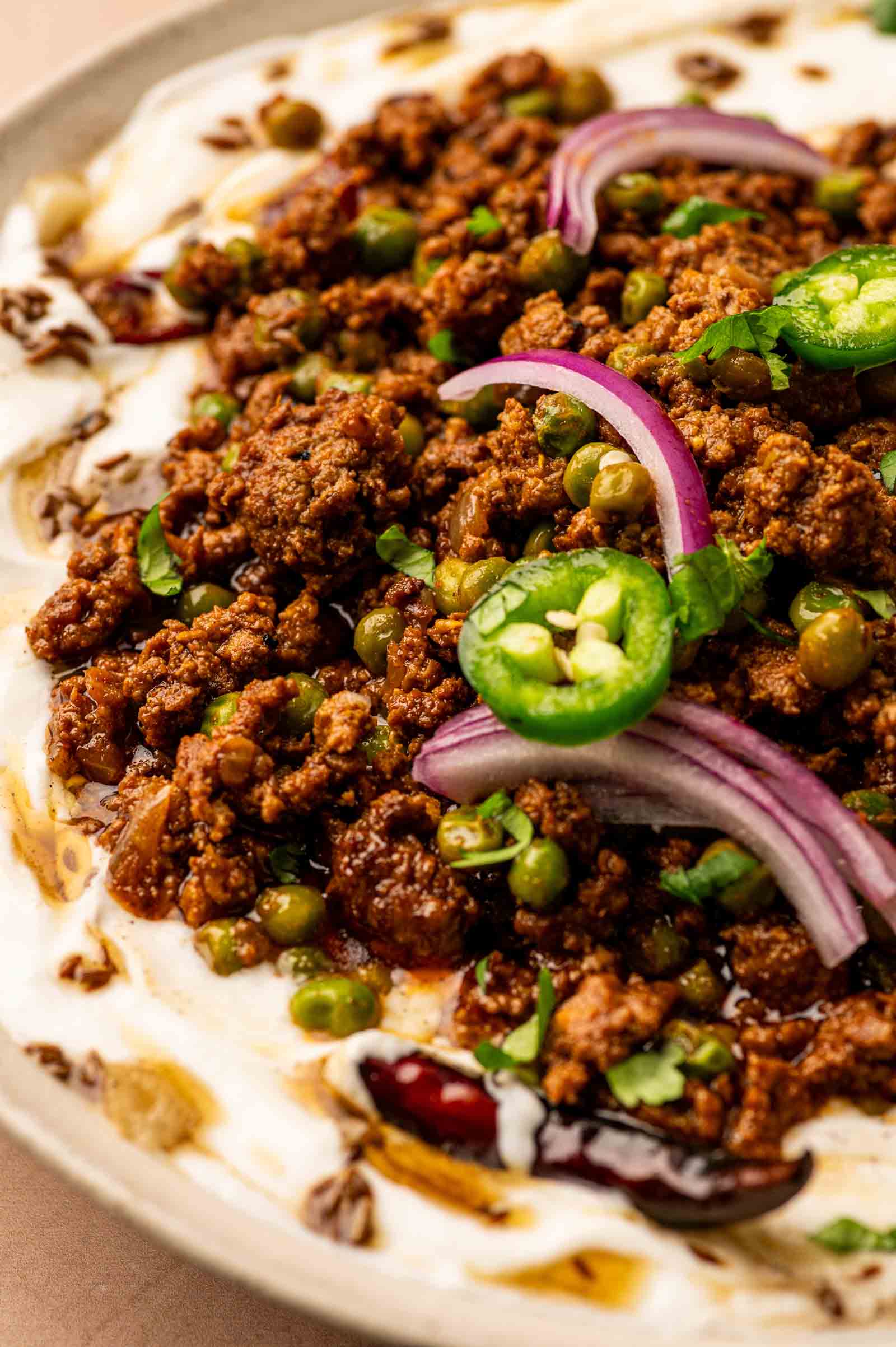 A close-up of a dish with spiced ground lamb keema, green peas, red onion slices, jalapeño, and herbs on a creamy white tadka swirled yogurt.