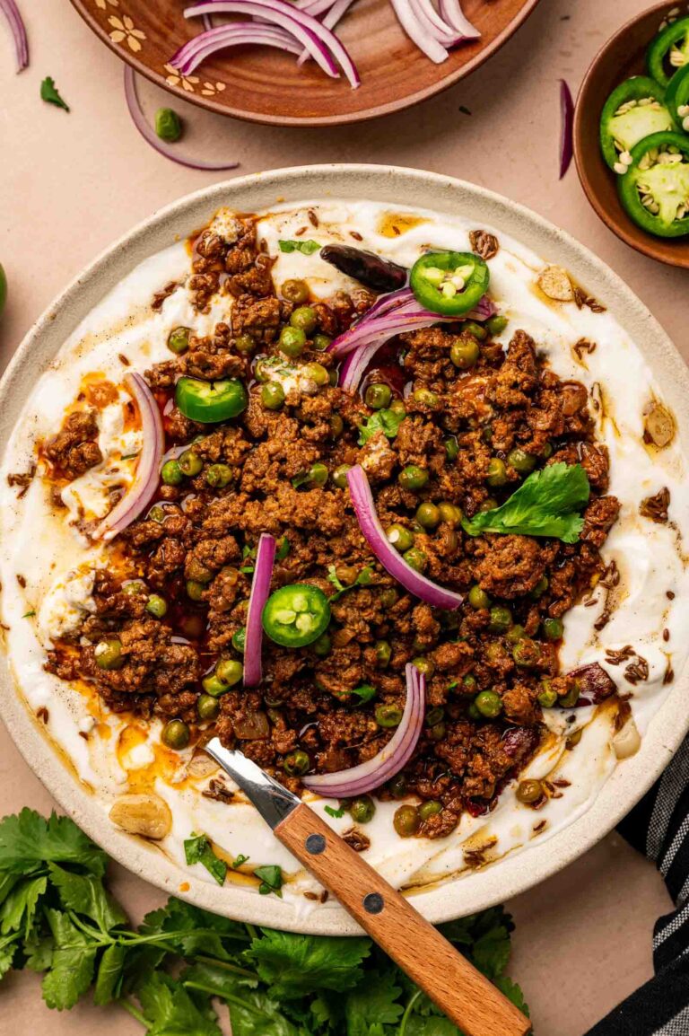 A plate of spiced ground lamb keema with peas, red onion, and jalapeño slices served over a creamy white sauce, with fresh herbs and a wooden-handled spoon on the side.