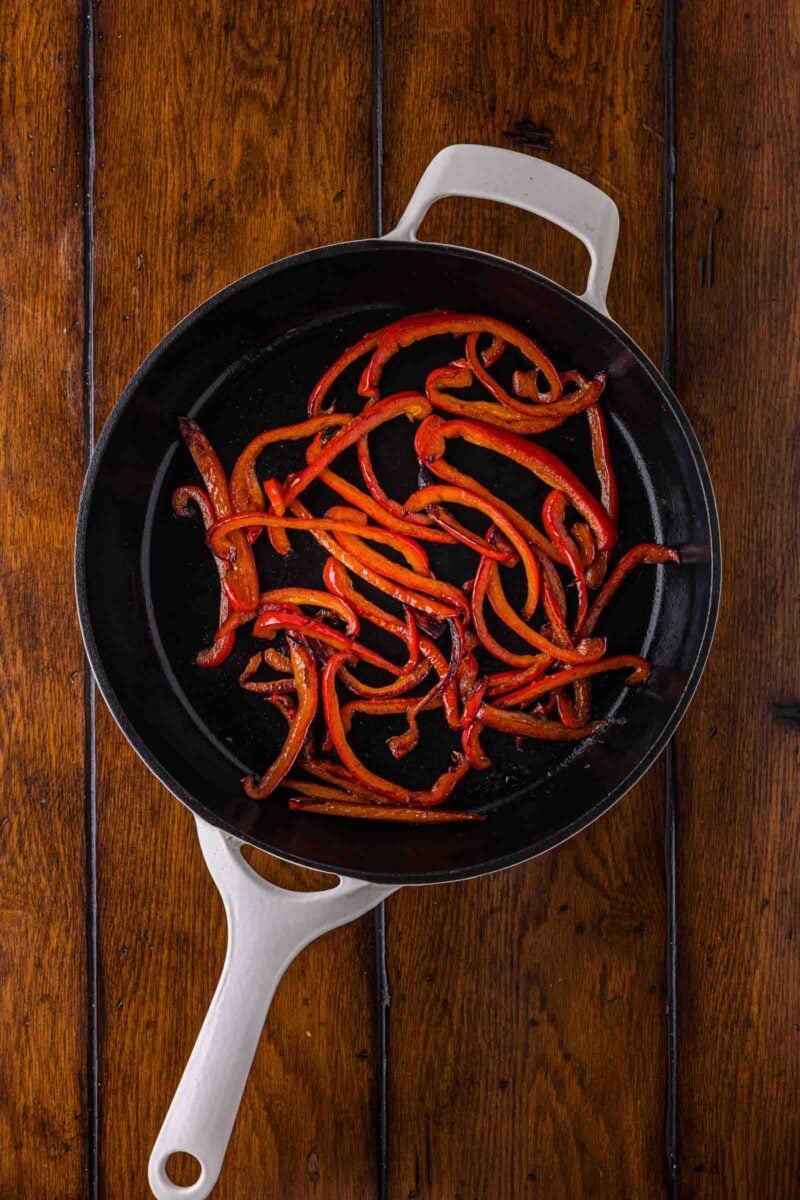 Sliced red bell peppers sautéed in a white-handled black skillet on a wooden surface.