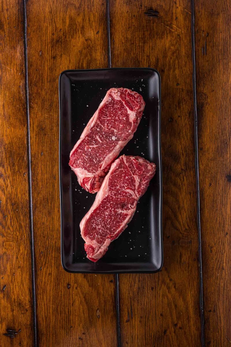 Two raw, marbled strip steaks with coarse salt on a black rectangular plate, placed on a wooden table.