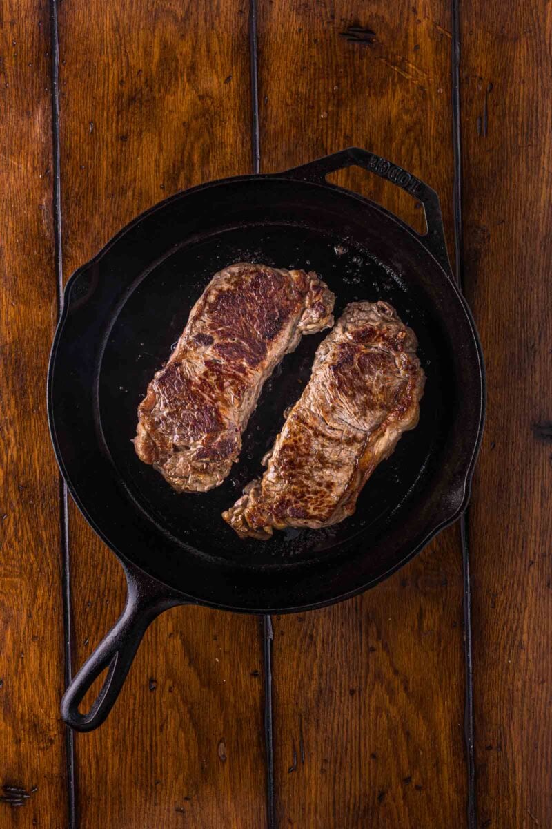Two cooked steaks in a black cast iron skillet on a wooden surface, viewed from above.