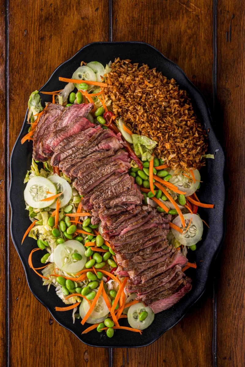A black plate with sliced steak on mixed salad with cucumbers, carrots, edamame, and a side of crispy rice, set on a wooden table.