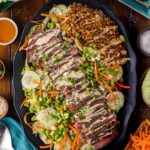 Platter with sliced steak drizzled with Japaense white sauce, shredded carrots, cucumber, edamame, lettuce, and a serving of crispy rice, surrounded by small bowls of sauces and vegetables.
