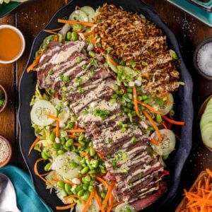 Platter with sliced steak drizzled with Japaense white sauce, shredded carrots, cucumber, edamame, lettuce, and a serving of crispy rice, surrounded by small bowls of sauces and vegetables.