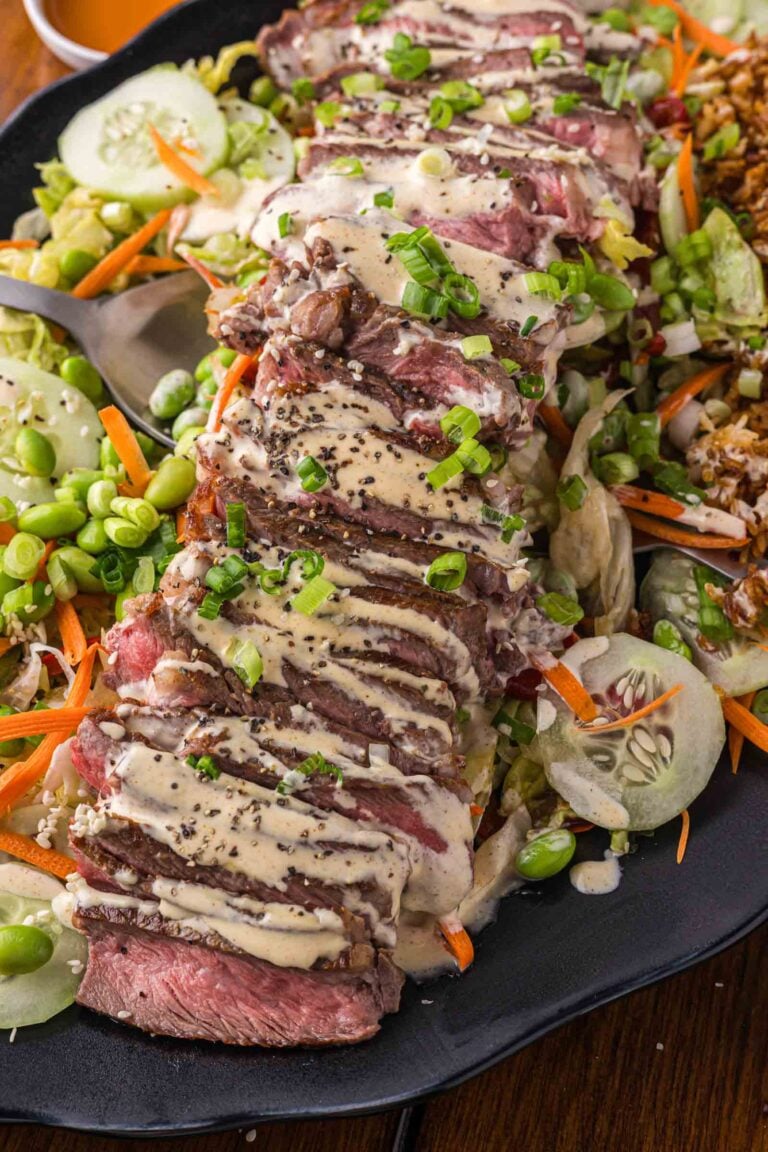 Sliced steak topped with Yum Yum sauce and chopped green onions, served over mixed salad with cucumbers, carrots, edamame, and crispy rice on a black platter.