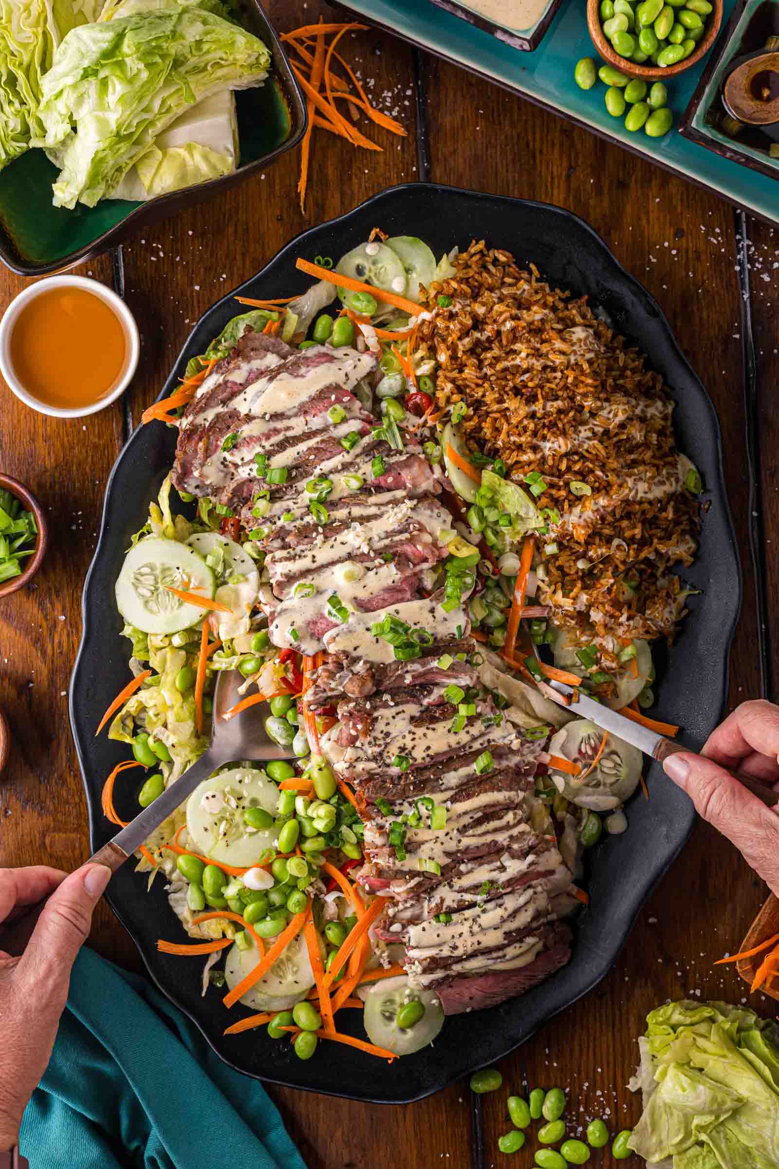 A platter with sliced steak drizzled with Japanese White Sauce, served with crispy rice, cucumbers, carrots, edamame, and chopped green onions. Hands hold serving utensils at the edges of the dish.