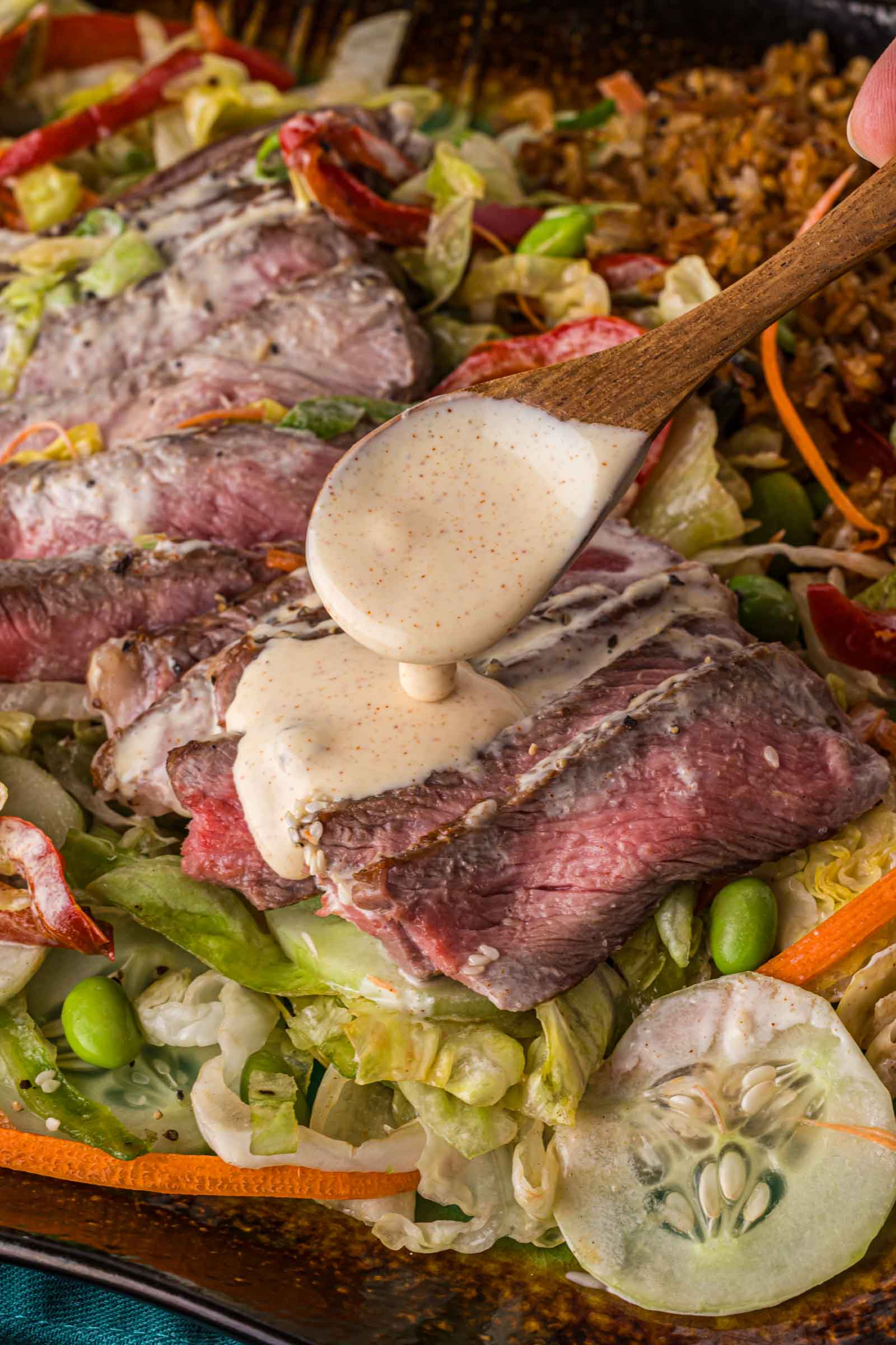 A spoon pours creamy dressing over sliced seared steak on a salad with lettuce, cucumber, carrots, edamame, and rice.