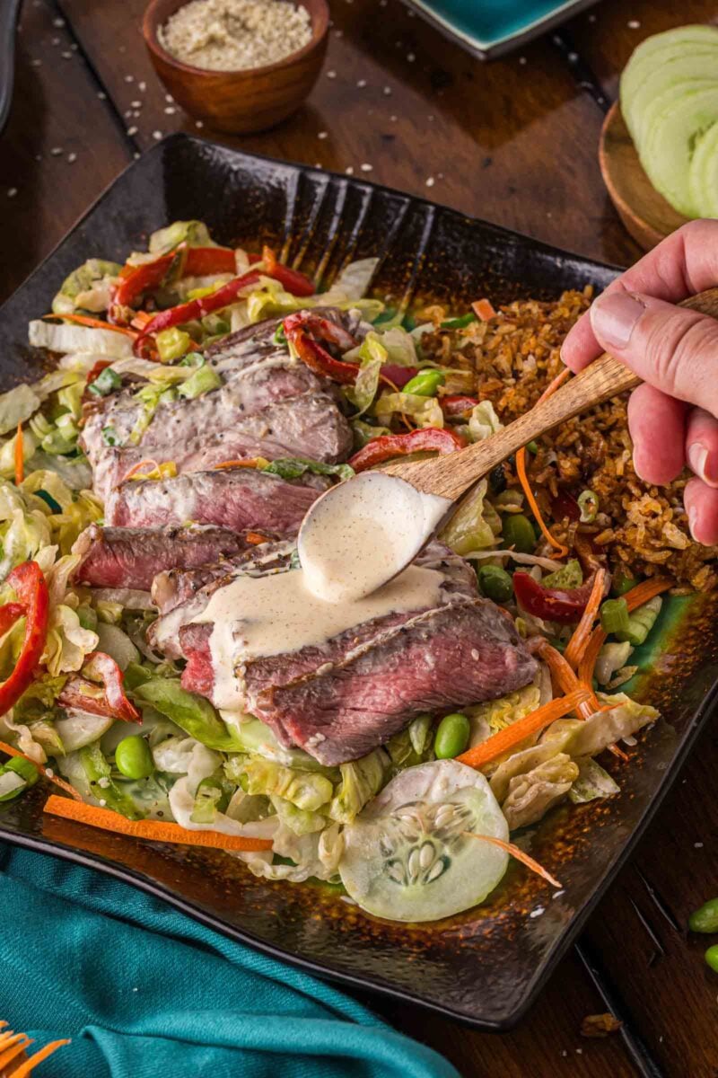A hand spoons creamy Yum Yum Sauce over Hibachi steak salad on a black plate.