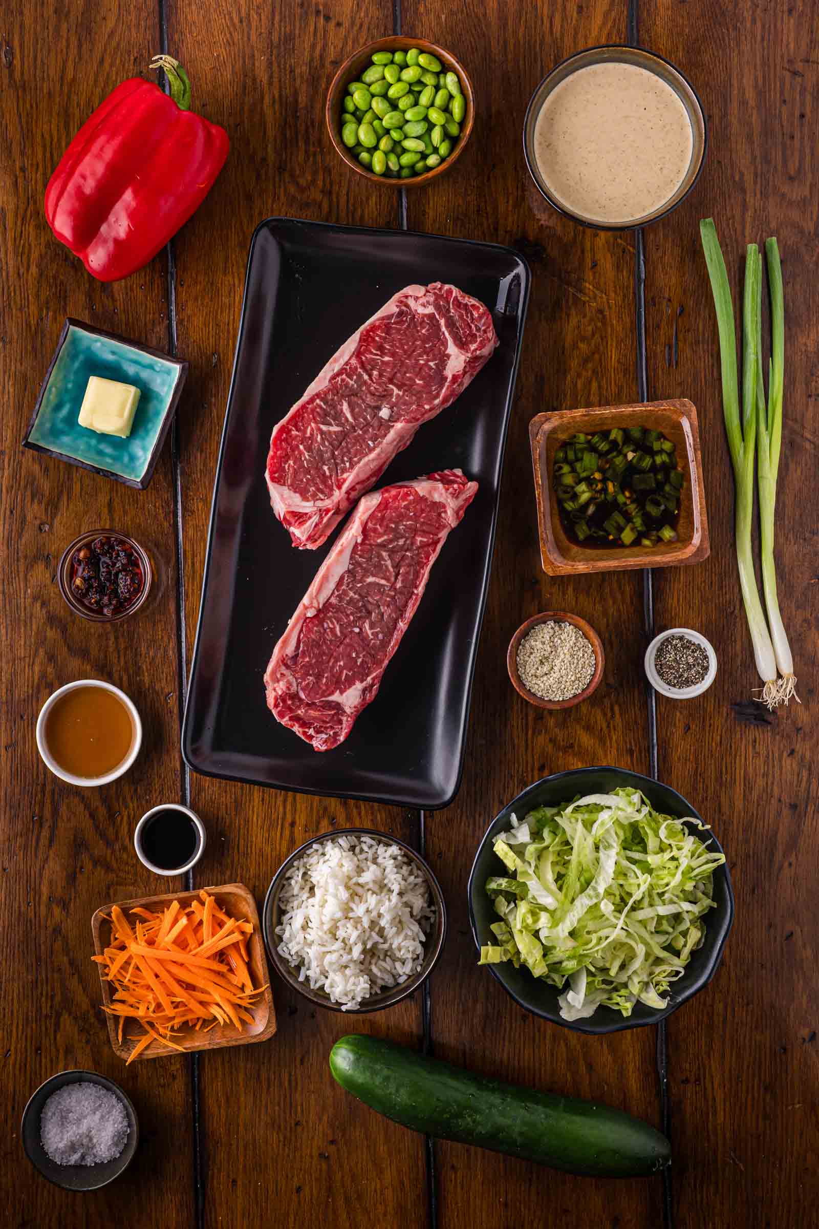 Overhead view of ingredients for hibachi steak salad: raw steak, rice, lettuce, carrot, cucumber, red pepper, edamame, spring onions, sauces, seasoning, butter, and other ingredients arranged on a wooden table.