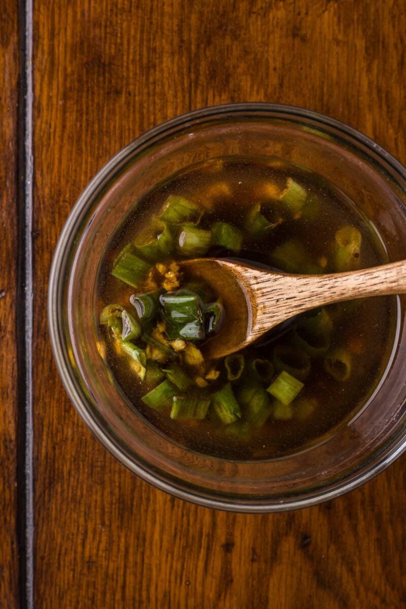 A glass bowl of Ginger Dressing with chopped green onions and a wooden spoon, placed on a wooden surface.