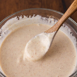 A glass bowl filled with a creamy, speckled yum yum sauce, with a wooden spoon resting inside, on a wooden surface.