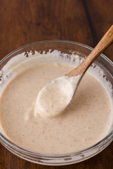 A glass bowl filled with a creamy, speckled yum yum sauce, with a wooden spoon resting inside, on a wooden surface.