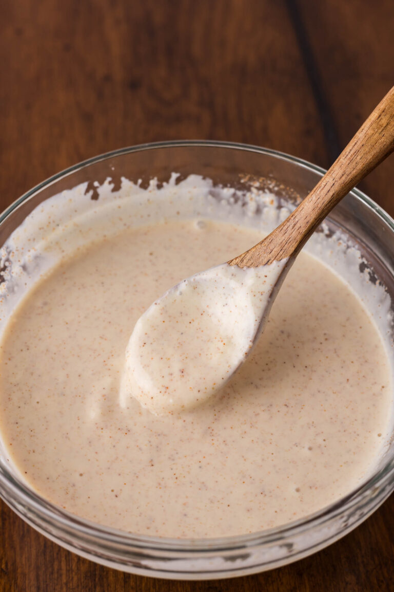 A glass bowl filled with a creamy, speckled yum yum sauce, with a wooden spoon resting inside, on a wooden surface.