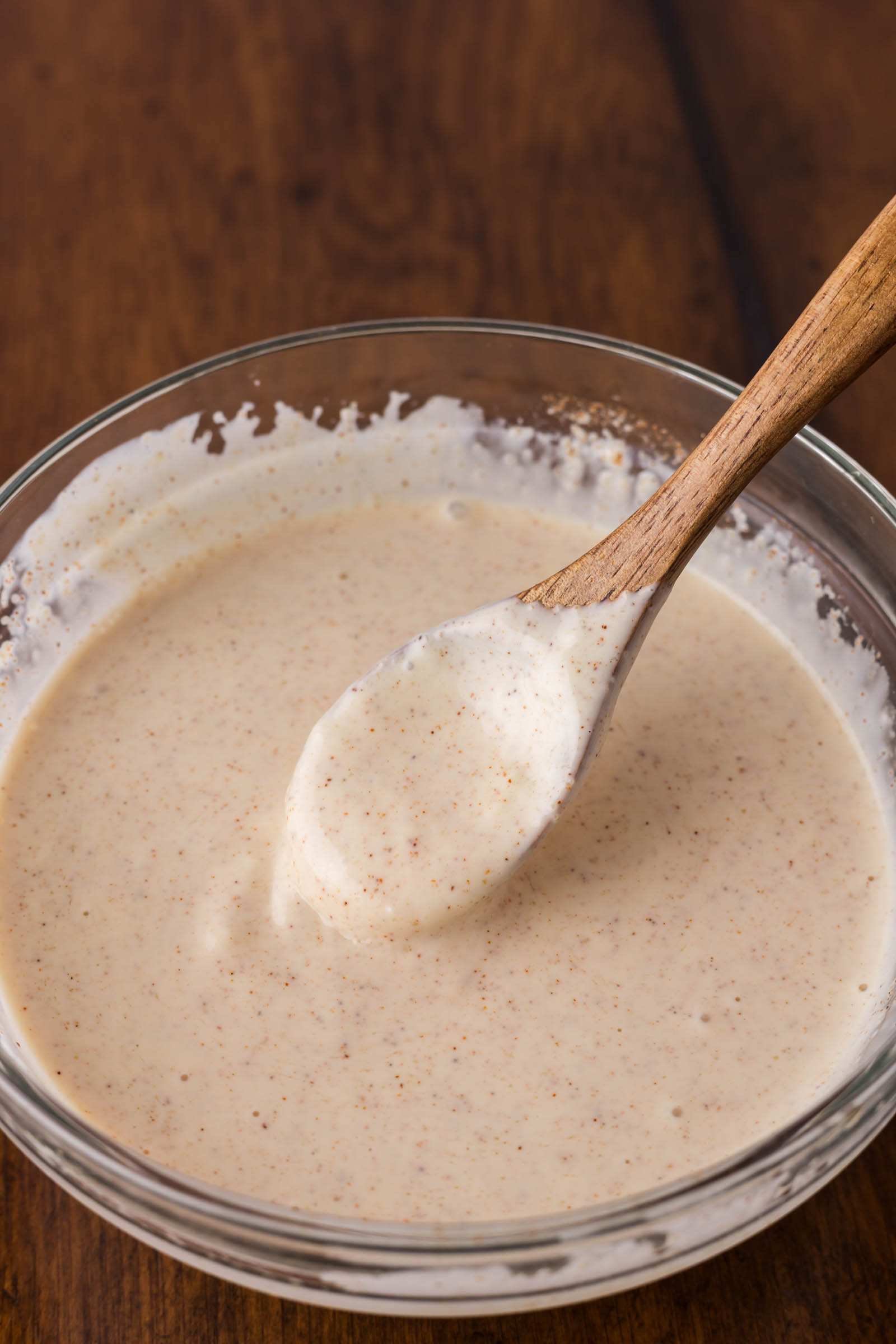 A glass bowl filled with a creamy, speckled yum yum sauce, with a wooden spoon resting inside, on a wooden surface.