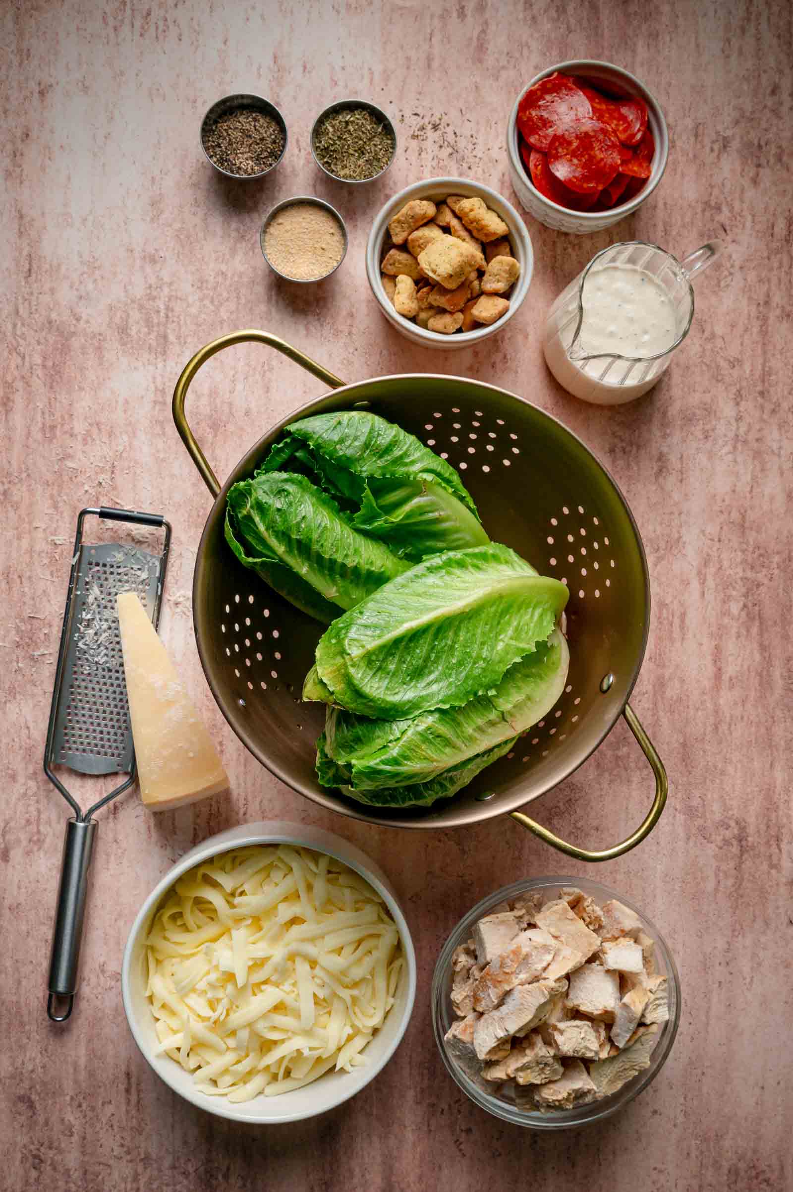 A colander with romaine lettuce, bowls of cooked chicken, shredded cheese, croutons, grated cheese, pepperoni, seasonings, and a cup of dressing are arranged on a countertop.