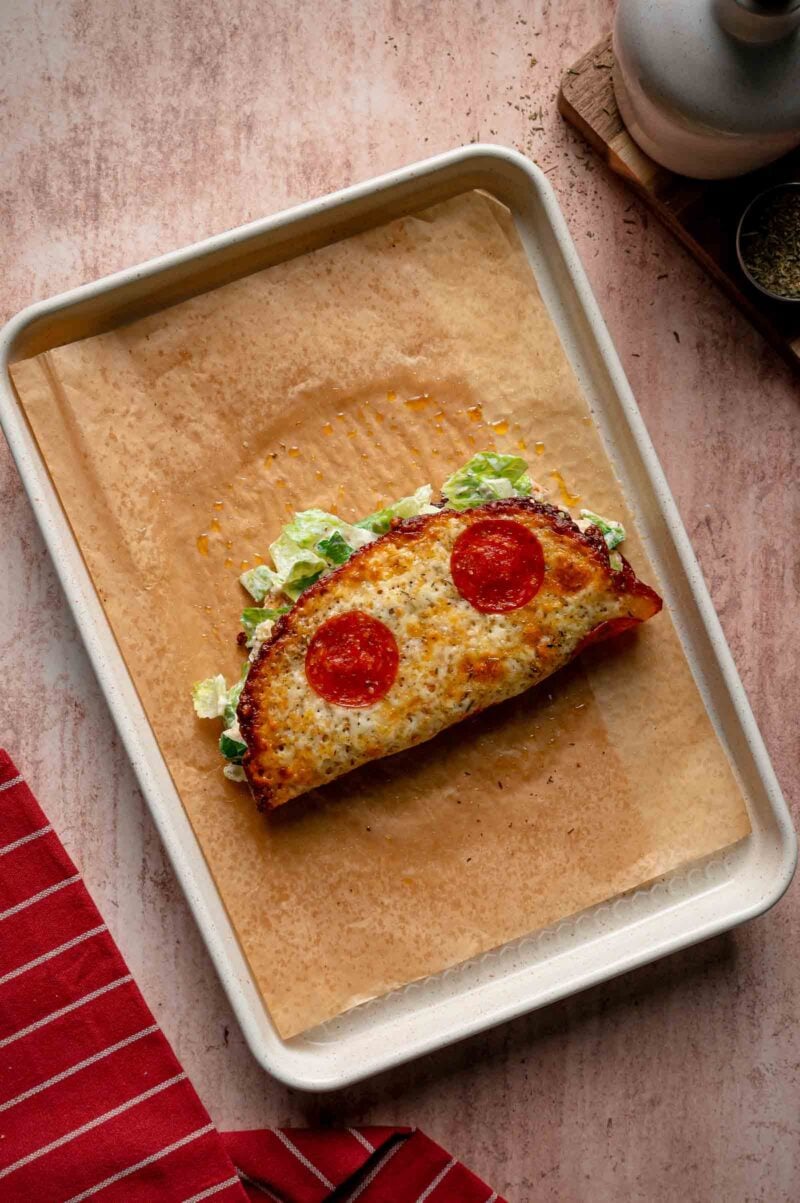 A pepperoni pizza caesar salad wrap is folded in half on a parchment-lined baking tray, with lettuce visible inside.