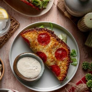 A pepperoni pizza Caesar salad wrap with a small bowl of extra dressing on the side, surrounded by various table items and a glass of lemon water.