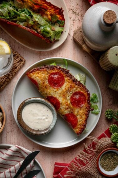 A pepperoni pizza Caesar salad wrap with a small bowl of extra dressing on the side, surrounded by various table items and a glass of lemon water.