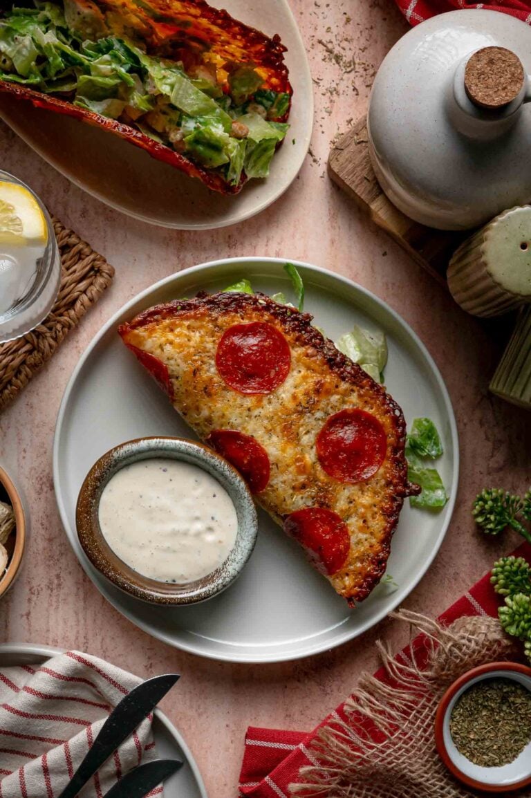 A pepperoni pizza Caesar salad wrap with a small bowl of extra dressing on the side, surrounded by various table items and a glass of lemon water.
