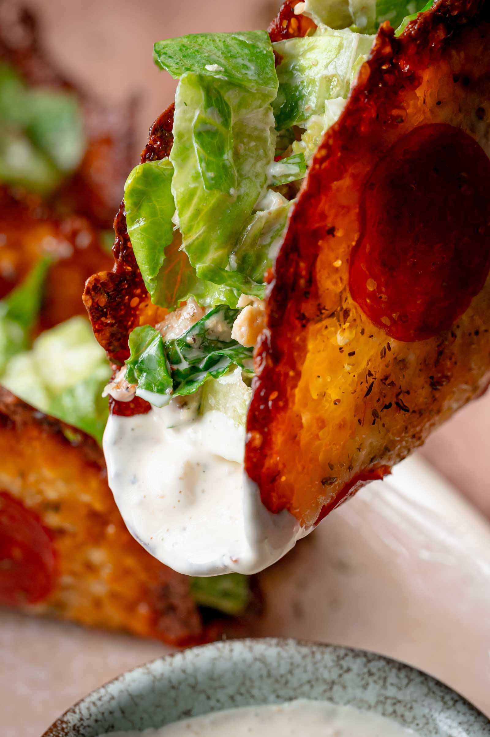 A shell made of melted cheese and pepperoni is filled with Caesar salad and dipped into a bowl of creamy Caesar dressing.