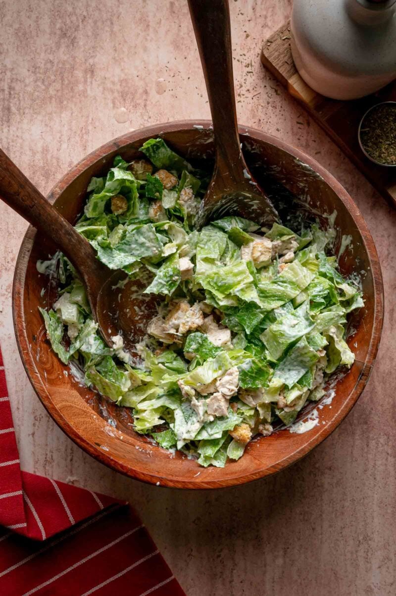Wooden bowl with Caesar salad, tossed with creamy dressing, croutons, and two wooden serving utensils on a beige surface.
