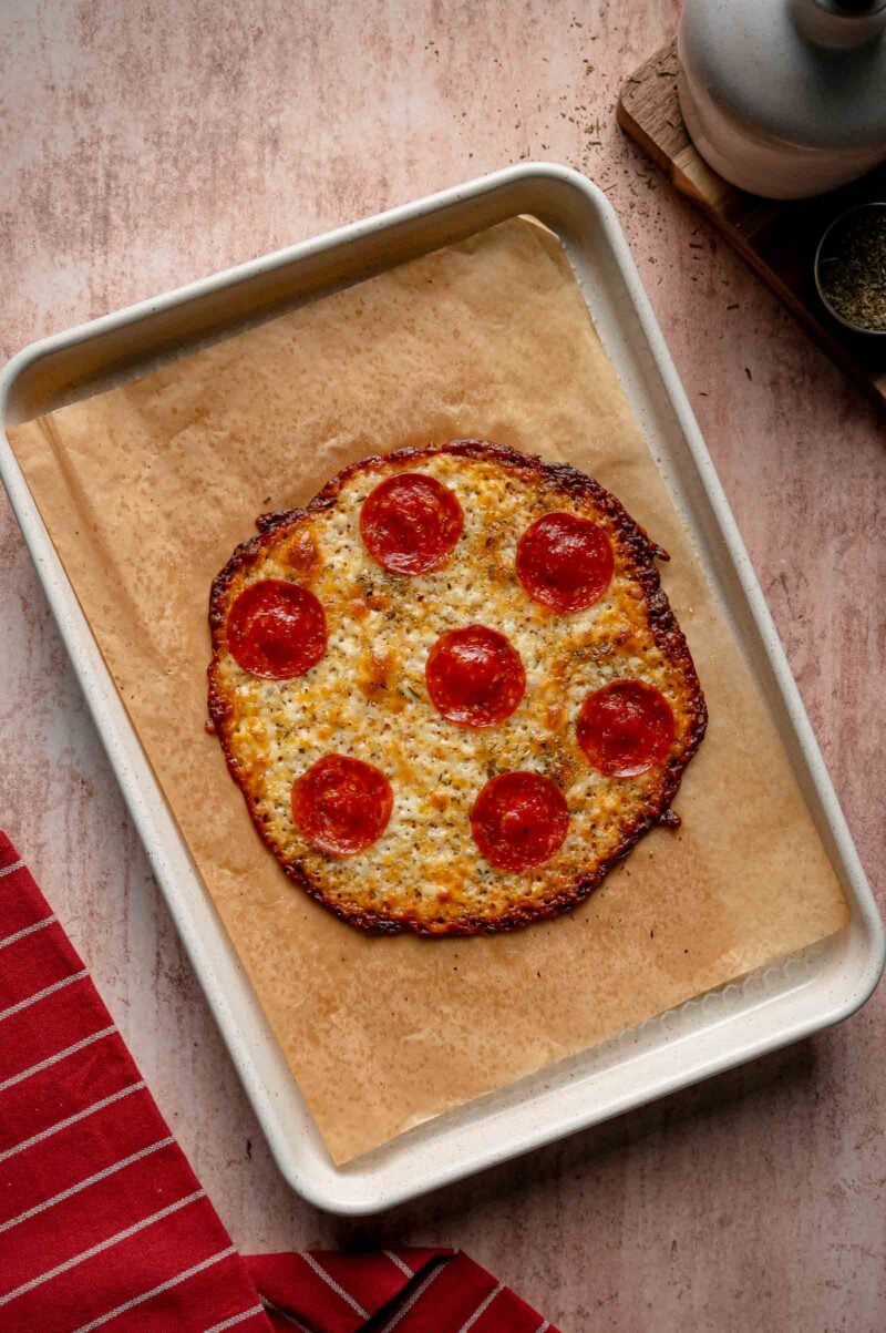 A round pepperoni pizza carb free wrap on parchment paper atop a baking sheet, next to a red-striped cloth and seasoning containers.