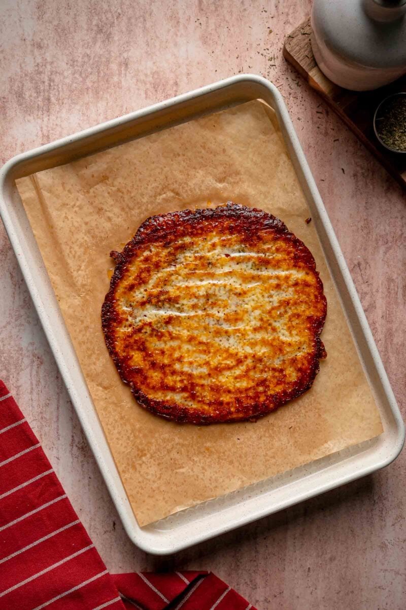 A baked round carb-free pizza wrap sits on parchment paper in a baking tray, with a red-striped cloth nearby.