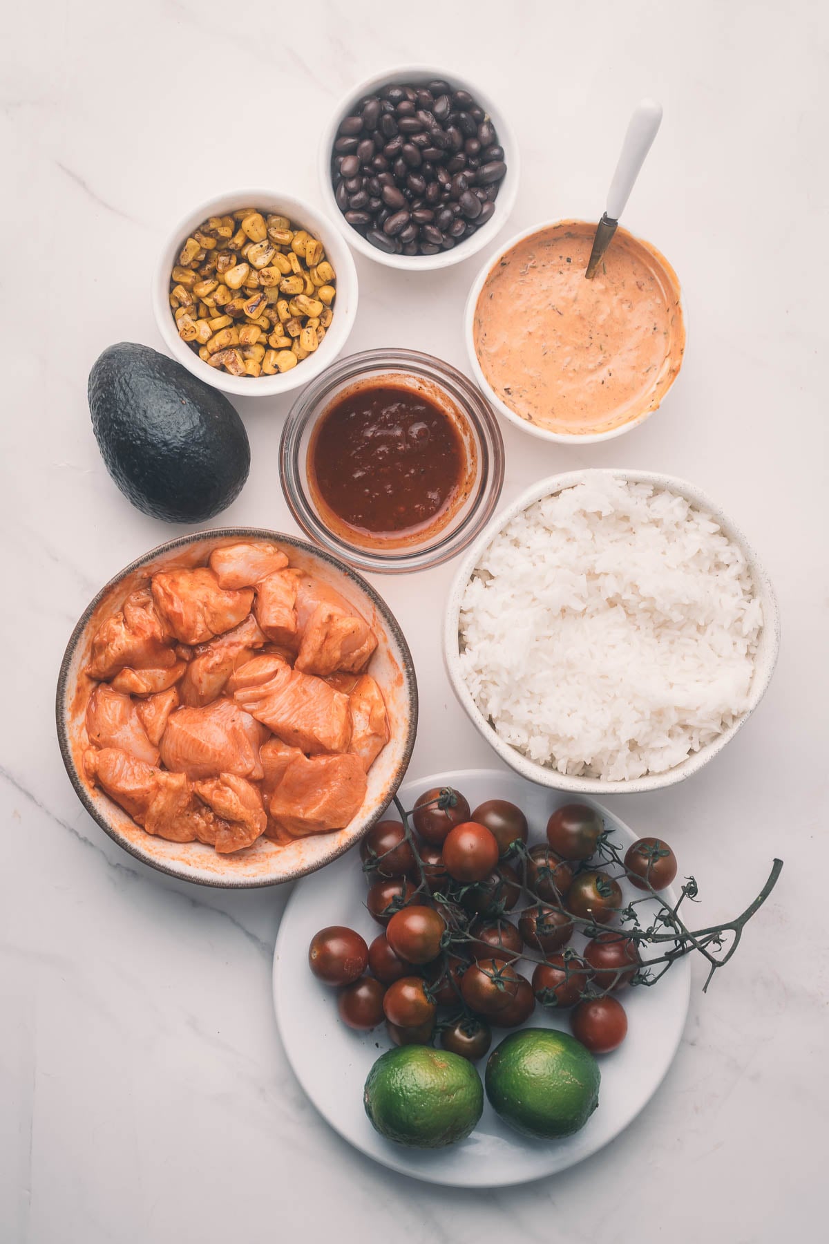 Bowls of marinated chipotle chicken, cooked rice, beans, corn, tomatoes, limes, avocado, sauce, and a creamy dressing are arranged on a white surface.