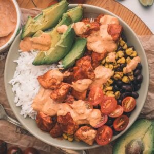 A bowl with white rice, bite-sized chipotle chicken, sliced avocado, cherry tomatoes, black beans, corn, and creamy sauce, garnished with lime wedges and surrounded by fresh ingredients.