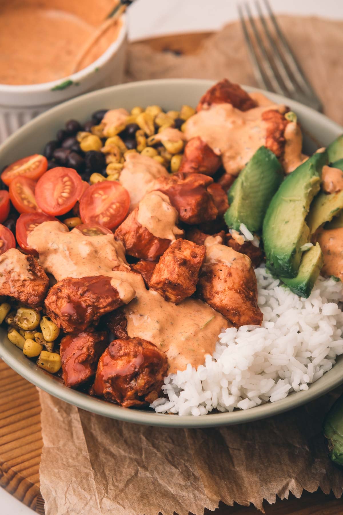 A bowl with white rice, chipotle chicken pieces, cherry tomatoes, black beans, corn, avocado slices, and creamy sauce, with a fork and sauce dish in the background.