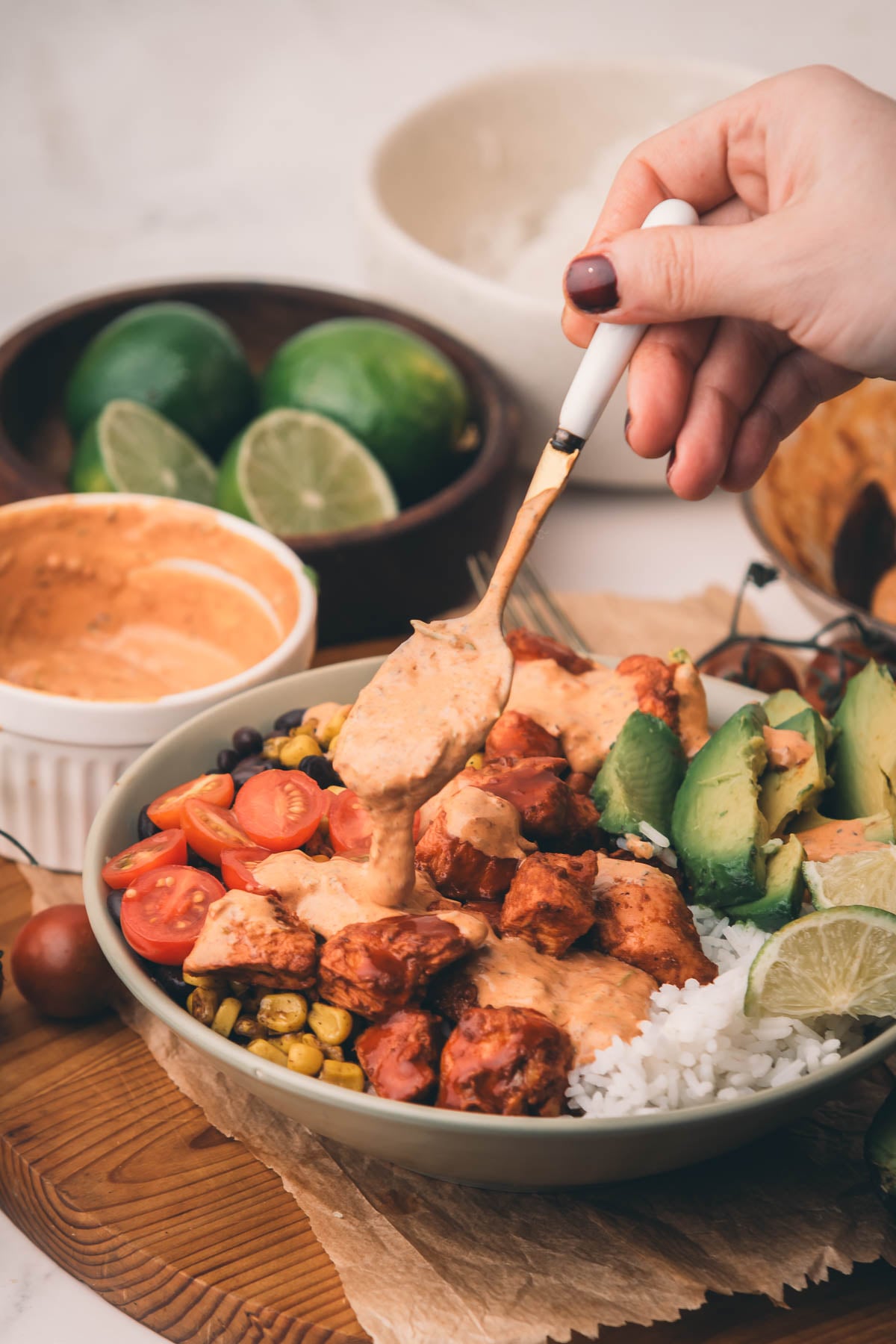 A hand drizzles sauce over a bowl with rice, chipotle chicken, cherry tomatoes, corn, black beans, avocado, and lime wedges.