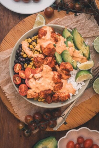 A bowl with rice, black beans, corn, cherry tomatoes, avocado slices, and chipotle chicken topped with a creamy sauce, garnished with lime wedges.