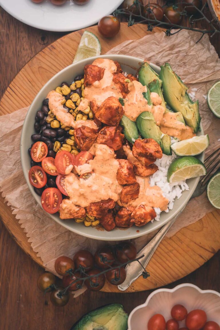A bowl with rice, black beans, corn, cherry tomatoes, avocado slices, and chipotle chicken topped with a creamy sauce, garnished with lime wedges.