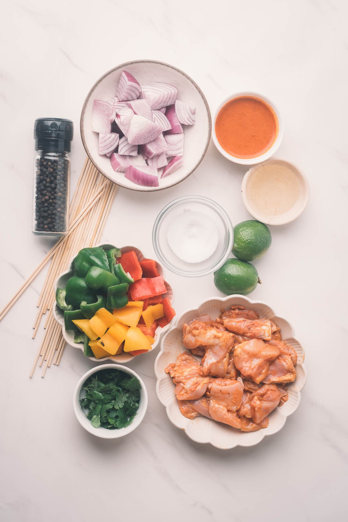 Ingredients for kebabs arranged on a table: chiptole marinated chicken, chopped onions, bell peppers, limes, cilantro, skewers, salt, pepper grinder, orange sauce, and a small bowl of oil.