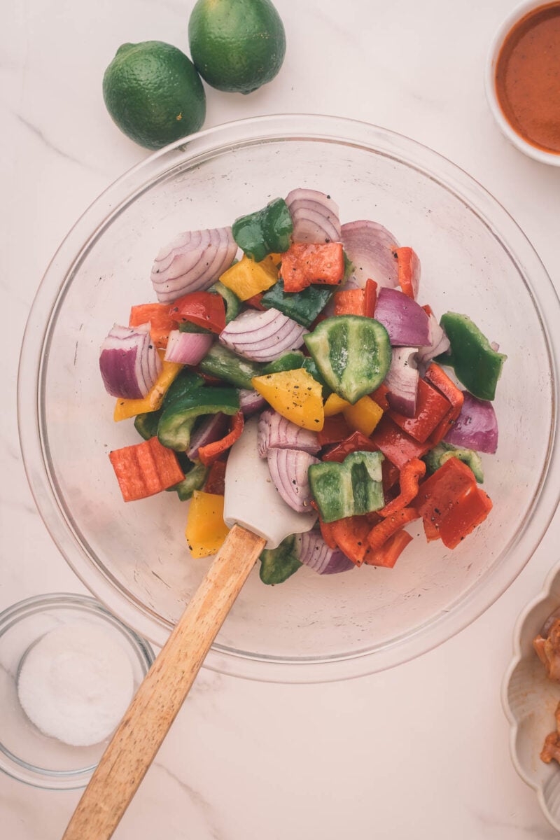 A glass bowl with chopped red onion, bell peppers, and cucumber being mixed with a spatula. Two limes, a small bowl of salt, and a sauce are nearby on a white surface.