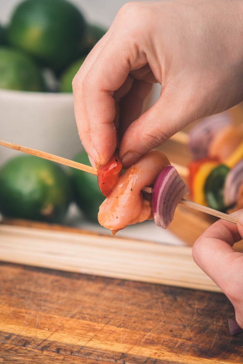 A hand threads marinated chicken, red onion, and red bell pepper onto a wooden skewer on a cutting board, with limes in the background.