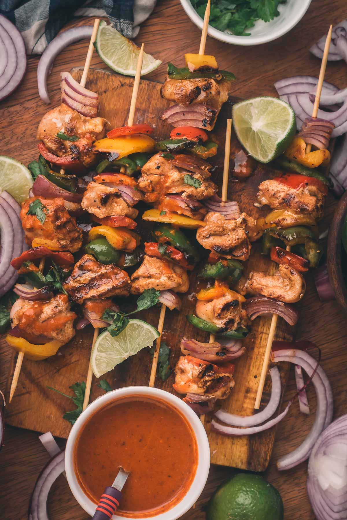 Grilled chiptole chicken and vegetable skewers on a wooden board, surrounded by lime wedges, red onion slices, cilantro, and a bowl of dipping sauce.