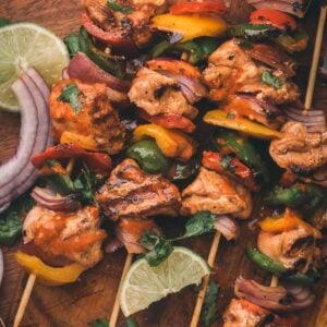 Grilled chipotle chicken and vegetable skewers on a wooden board, garnished with lime slices, red onion, and cilantro, with a bowl of sauce nearby.