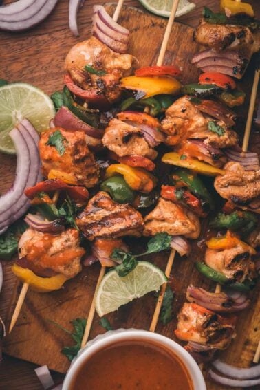 Grilled chipotle chicken and vegetable skewers on a wooden board, garnished with lime slices, red onion, and cilantro, with a bowl of sauce nearby.