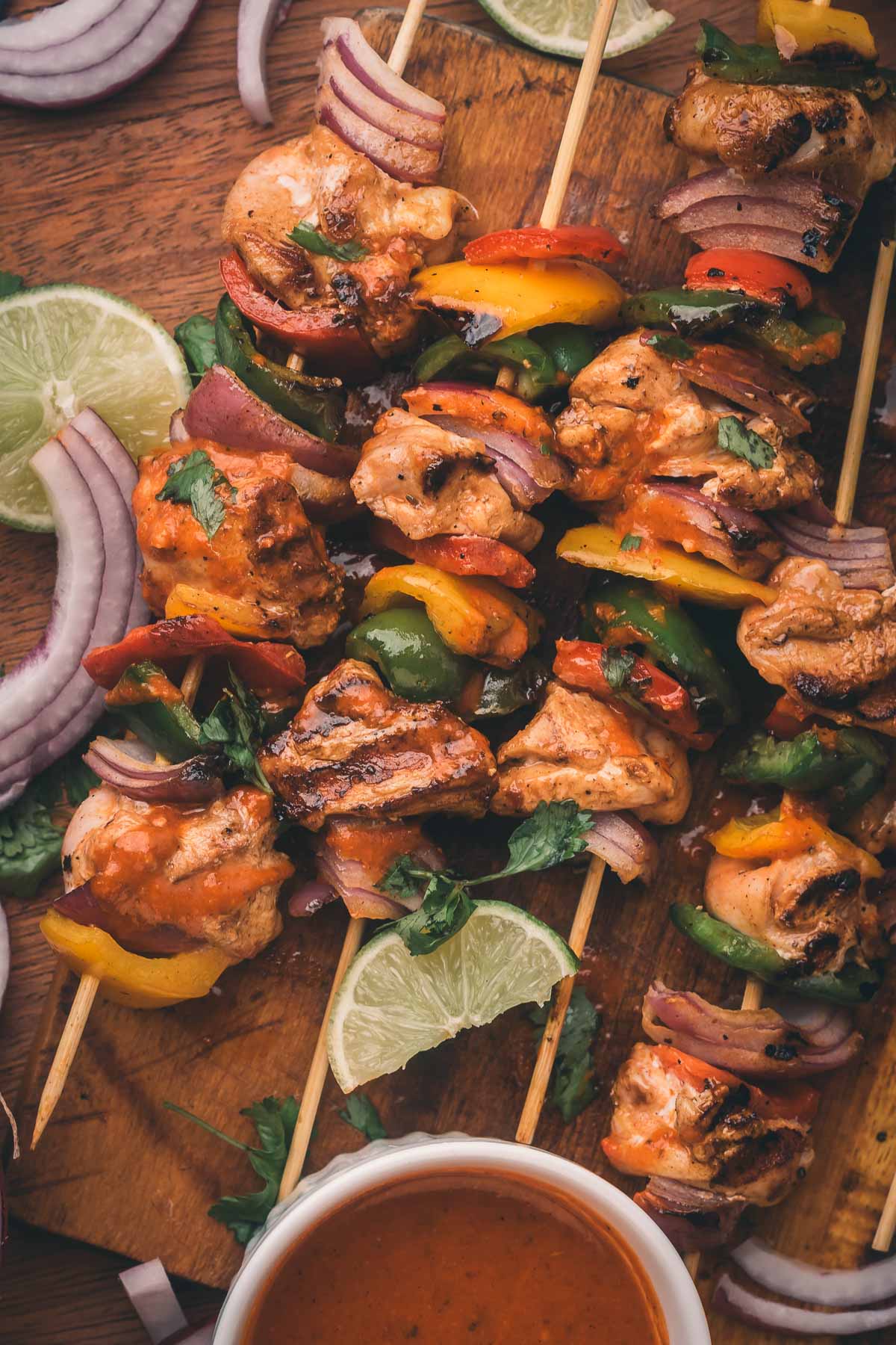Grilled chipotle chicken and vegetable skewers on a wooden board, garnished with lime slices, red onion, and cilantro, with a bowl of sauce nearby.
