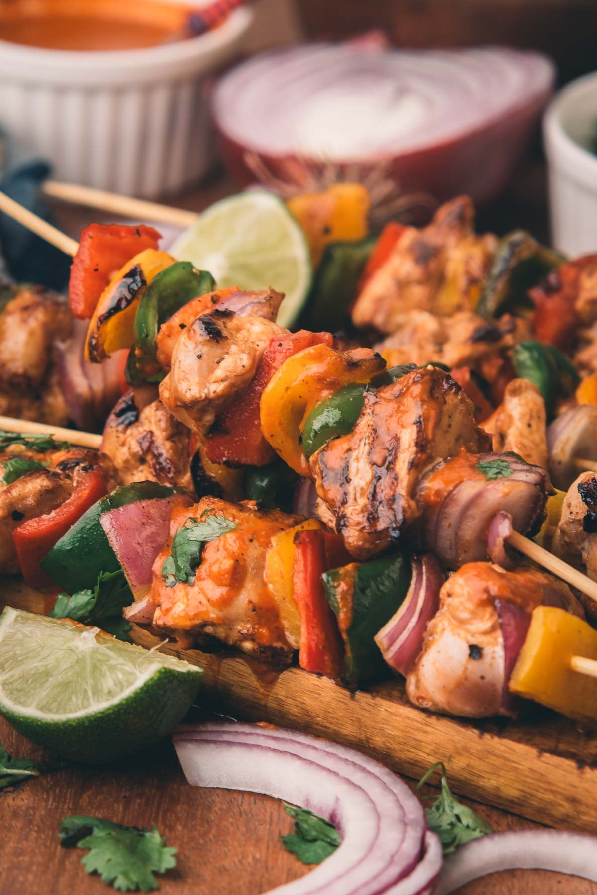 Grilled chipotle chicken skewers with bell peppers and red onions on a wooden board, garnished with lime wedges and cilantro, with dipping sauces in the background.
