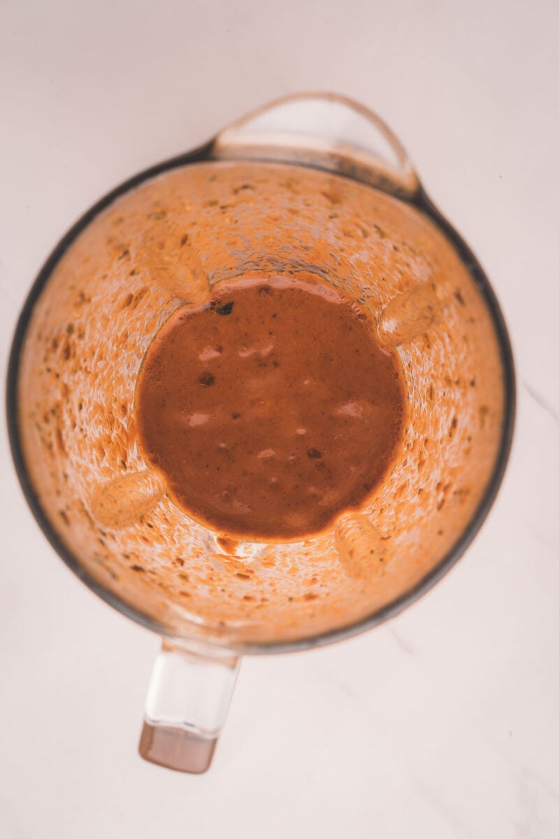 Overhead view of a blender containing a orange colored chipotle marinade with visible herb and spice flecks, resting on a white surface.
