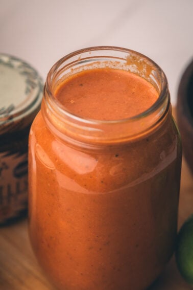 A glass jar filled with thick, orange-red sauce sits on a wooden surface next to a partially visible jar and a green lime.