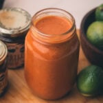 A glass jar filled with reddish-orange chipotle marinade sits on a wooden surface near a bowl of limes and two jars with sealed lids.
