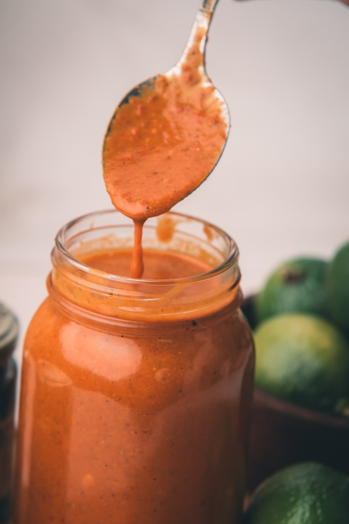A spoon drips chipotle marinade into a glass jar, with limes blurred in the background.