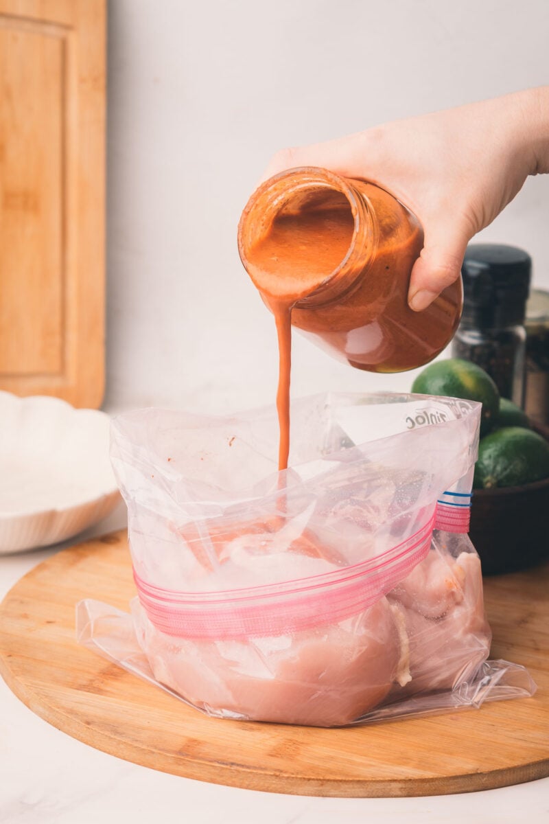 A hand pours chiptole marinade from a jar into a zip-top plastic bag containing raw chicken breasts on a wooden surface.