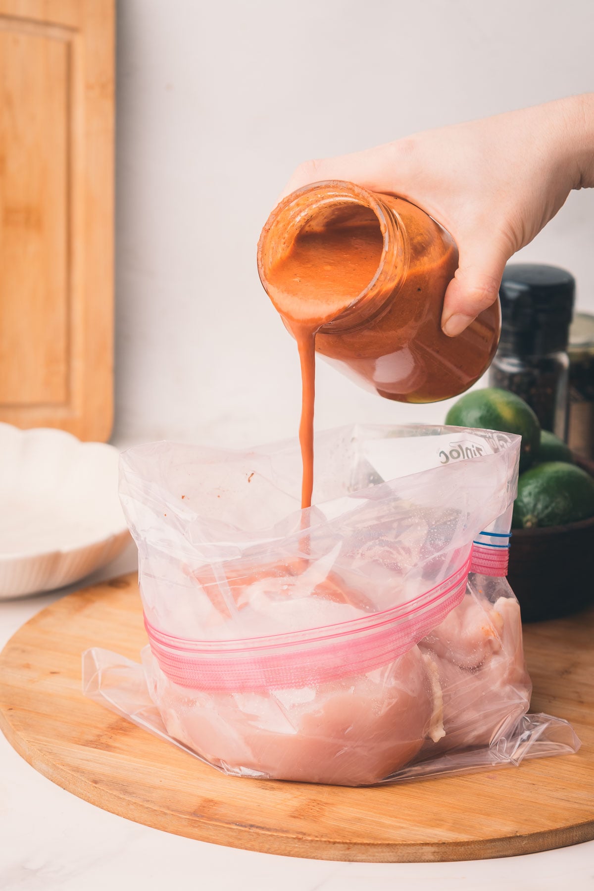 A hand pours chiptole marinade from a jar into a zip-top plastic bag containing raw chicken breasts on a wooden surface.