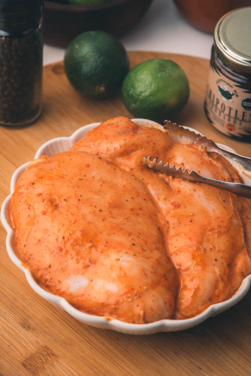 Two raw chicken breasts coated in chiptole marinade sit on a white plate with metal tongs, surrounded by limes, a spice jar, and a pepper grinder on a wooden surface.