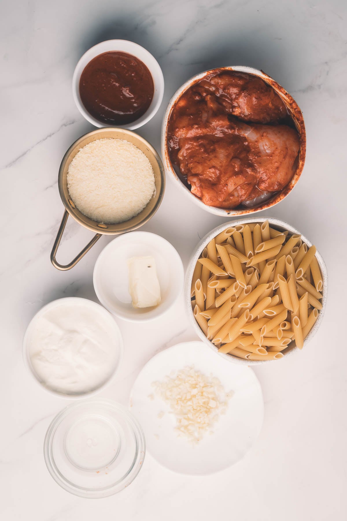 Top-down view of uncooked pasta, chipotle marinated chicken, grated cheese, sour cream, sauce, butter, and seasonings in separate bowls on a white surface.