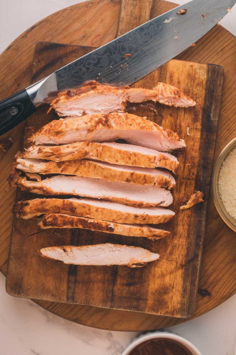 Sliced cooked chicken breast on a wooden cutting board, with a large knife resting beside it.